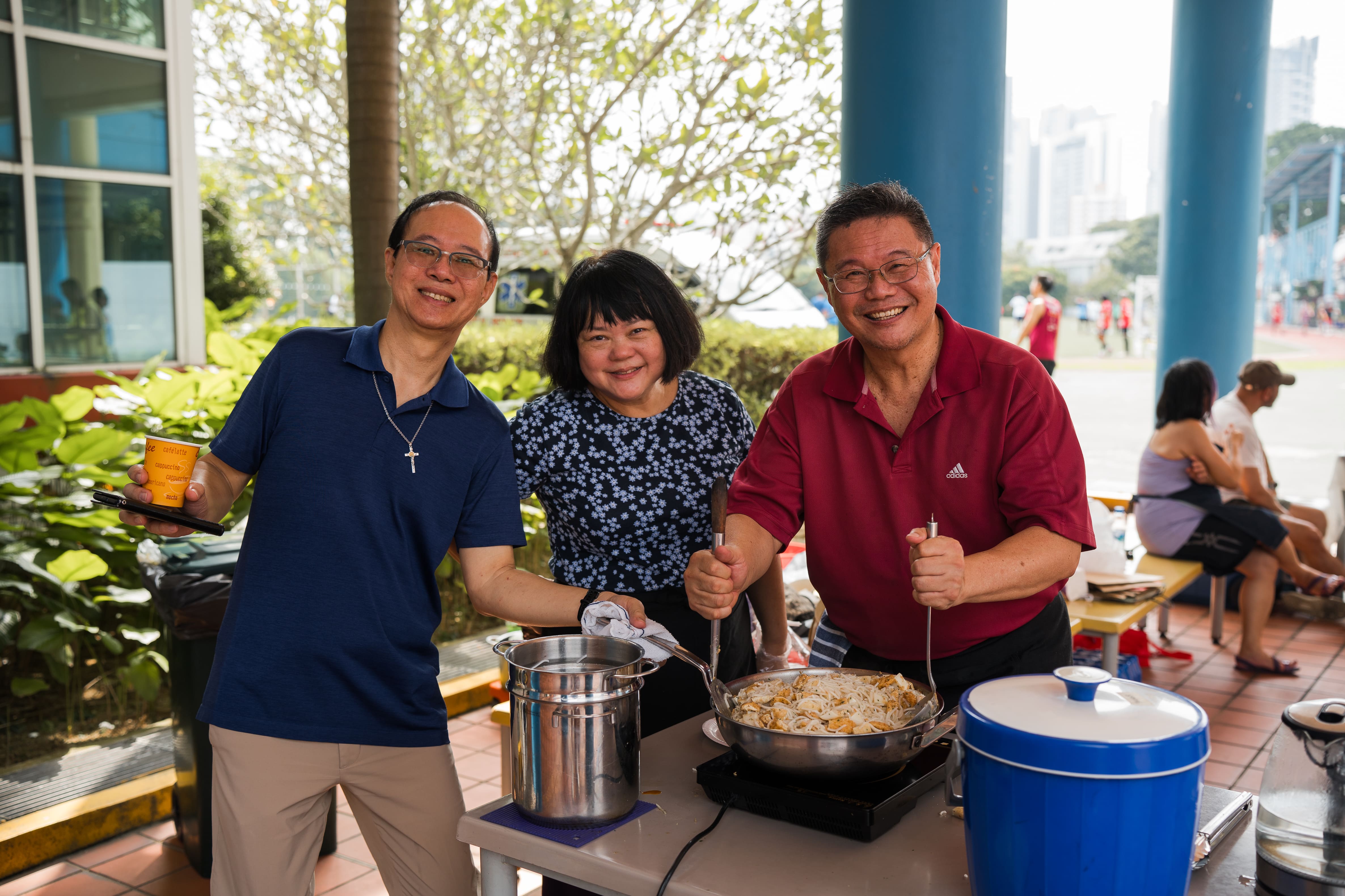Three smiling people stand at an outdoor table with a large wok of noodles and a pot on an induction stove.